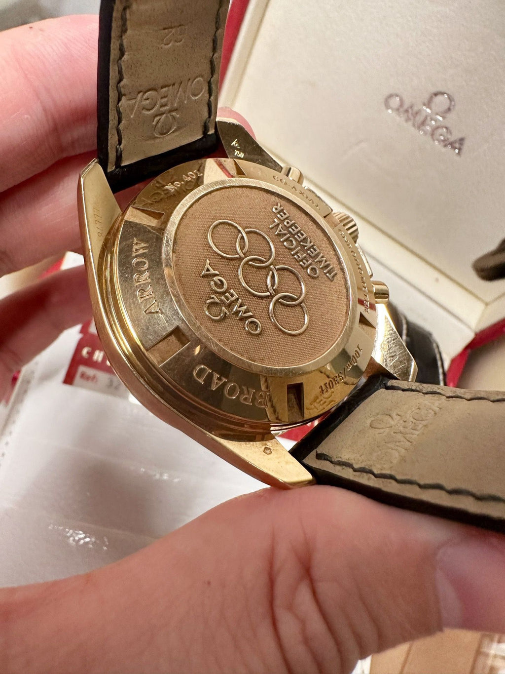 Gold watch case with Omega branding held by a hand, against a white background.