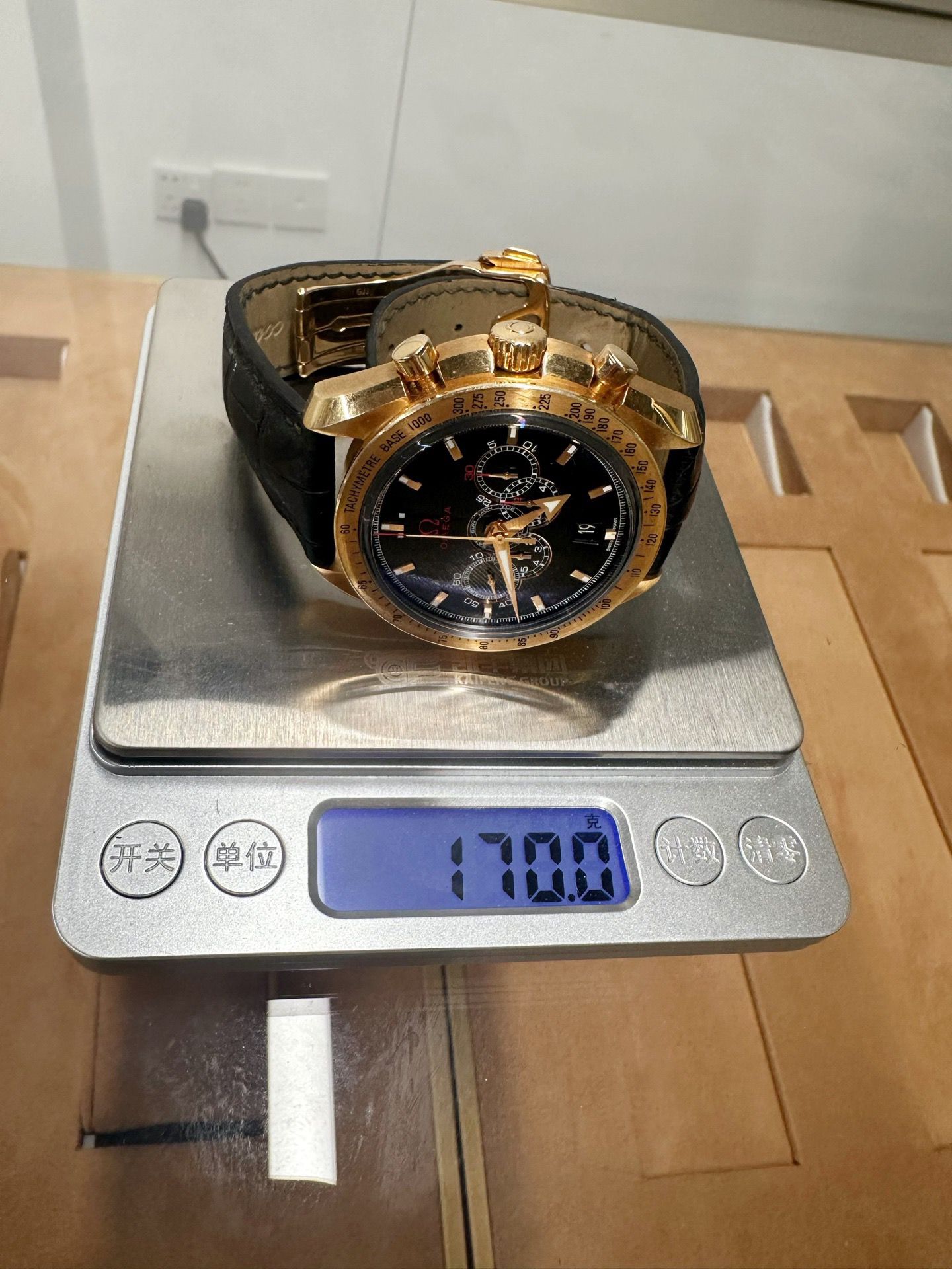 Omega Gold watch being weighed on a digital scale with a white wall in the background