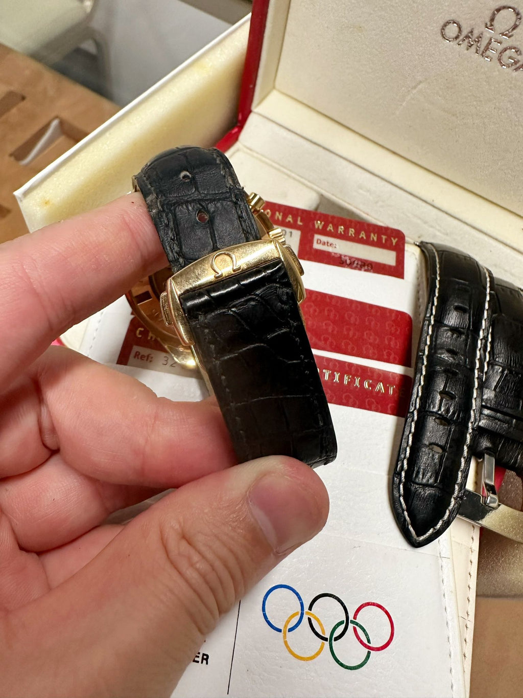 Black watch strap held by a hand with Omega branding, displayed against a background featuring Olympic rings and a warranty card.
