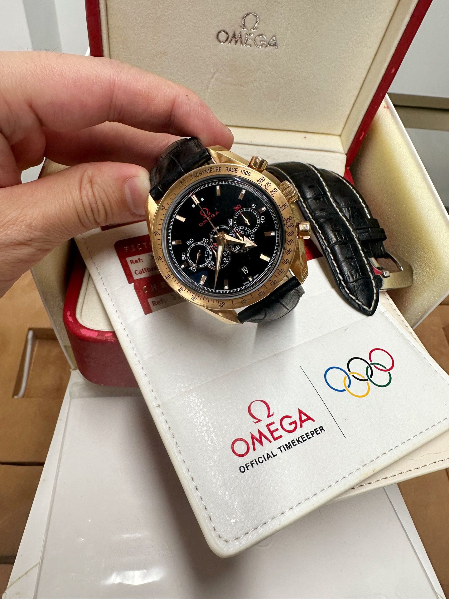 Gold watch with black leather strap held by a hand, displayed on an Omega branded card.