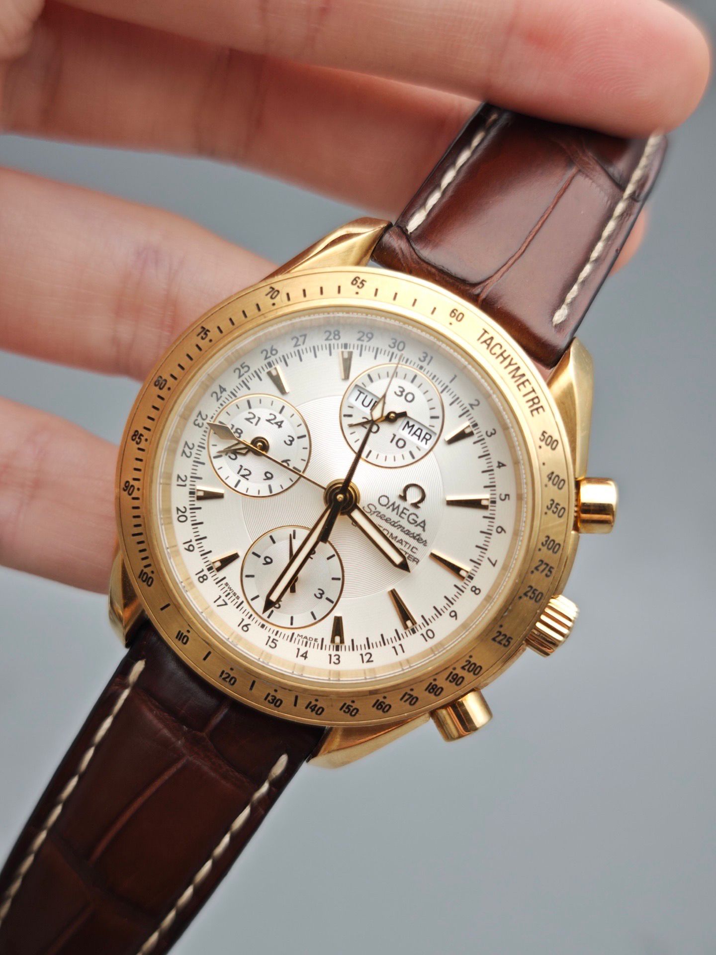Gold Omega watch with brown leather strap held by a hand against a gray background