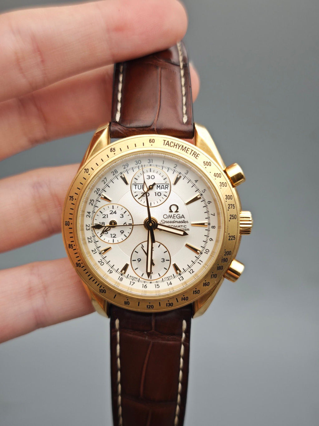 Gold Omega watch with brown leather strap held by a hand against a gray background