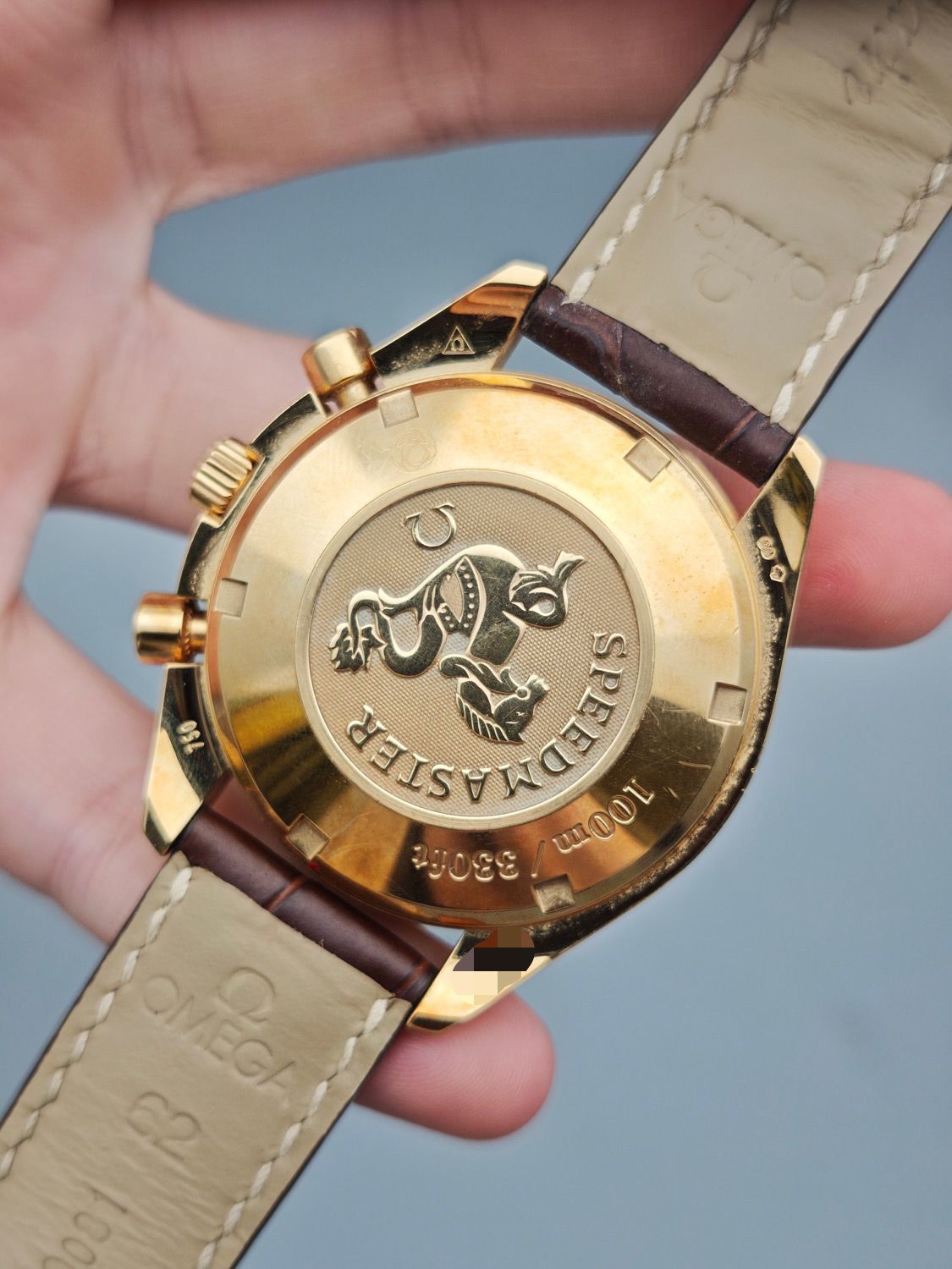 Gold Omega watch with a brand logo held by a hand against a blue background