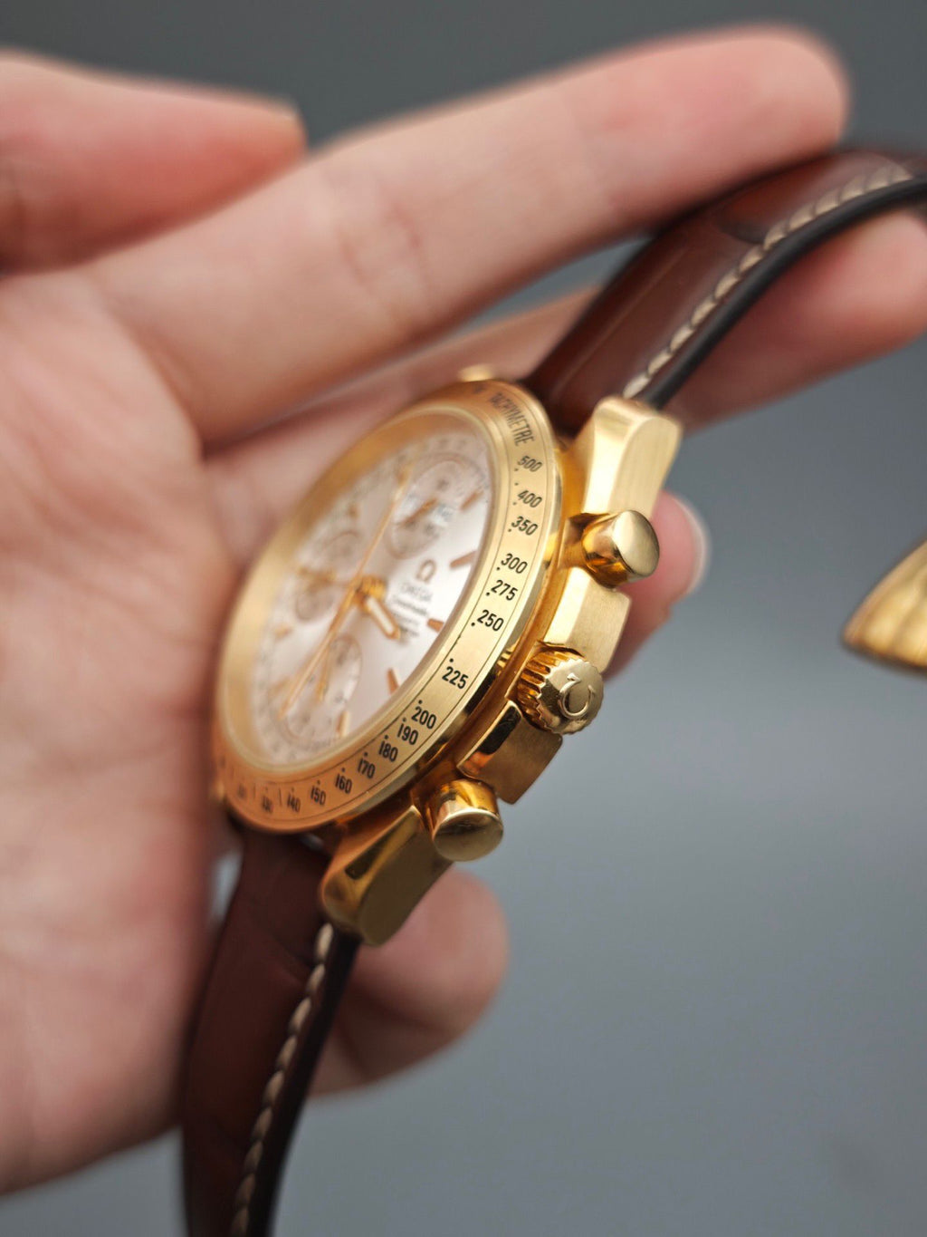 Gold Omega wristwatch with brown leather strap held in a hand against a gray background